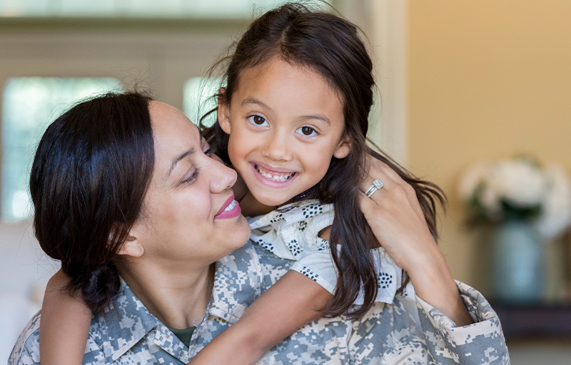 Adult in military uniform holding a child in a home setting.