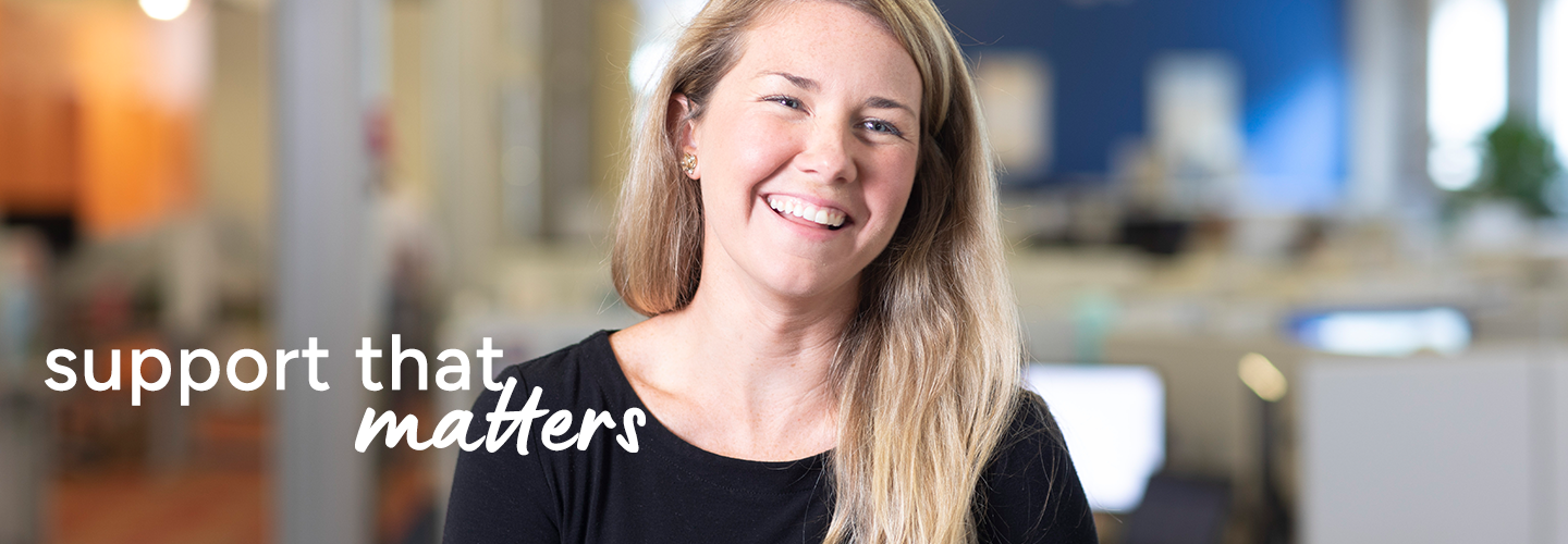 Smiling professional in a bright office with desks and computers, promoting supportive workplace culture.