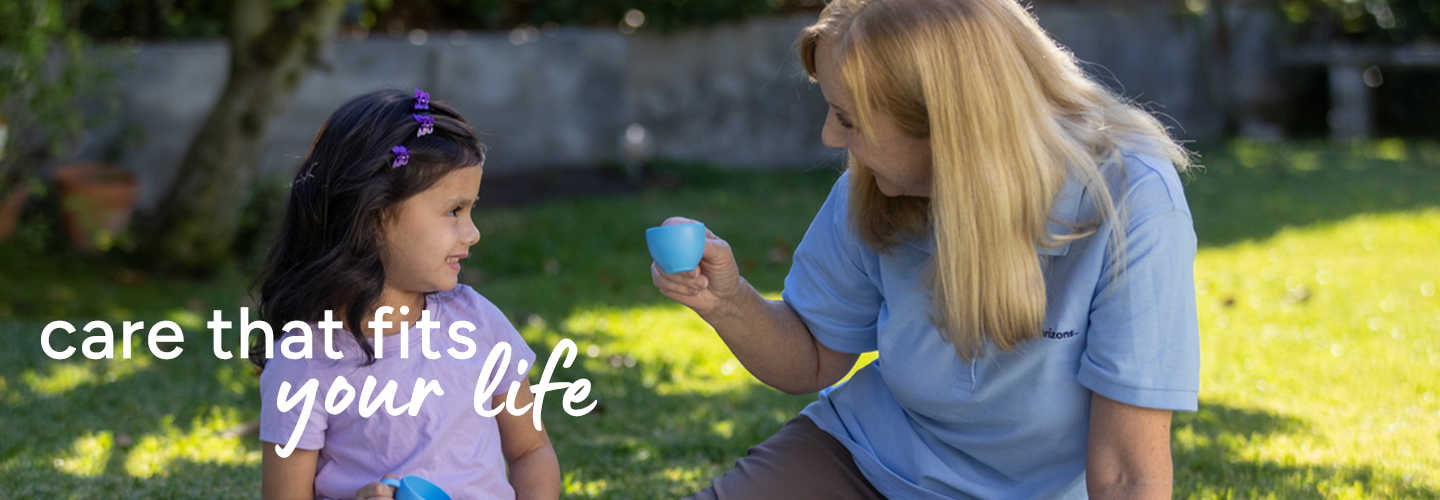 Two people sitting on grass outdoors, holding small blue cups as if sharing a pretend tea party. Text overlay reads “care that fits your life.
