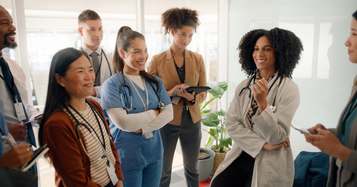 Diverse healthcare team in hallway, smiling and talking. Some wear scrubs or lab coats, others hold tablets or notebooks.
