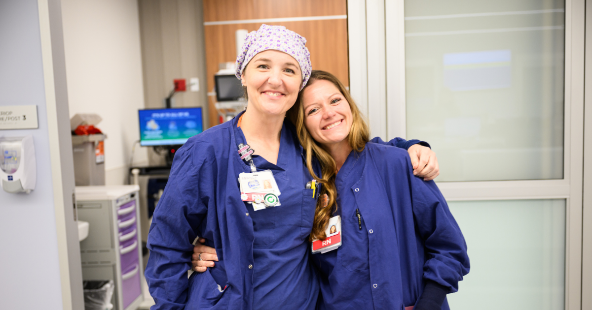 Smiling female healthcare professional with short white hair wearing navy blue scrubs and a stethoscope, standing confidently with arms crossed in a clinical setting.