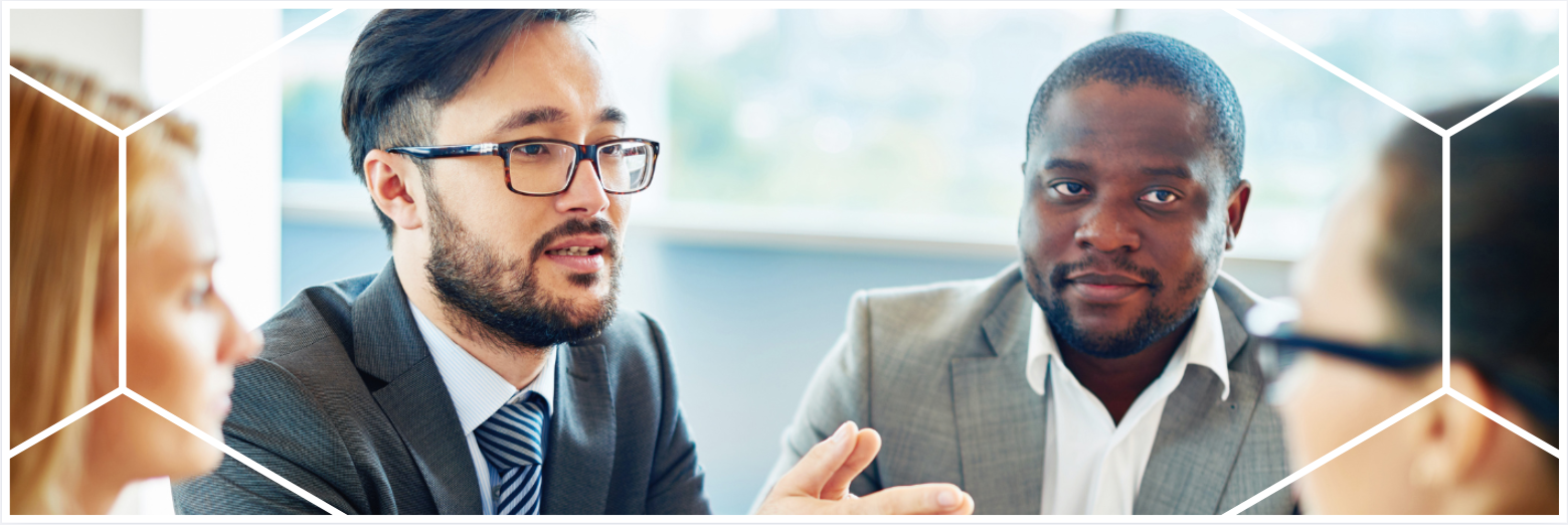 A group of professionals in business attire sit at a table in discussion. One man gestures as he speaks, while another looks attentively at a colleague. The image is framed with a white geometric brand overlay.
