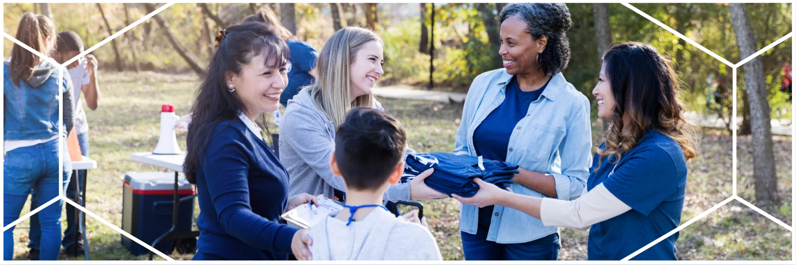 Four women and one child engage in a cheerful outdoor volunteer event, handing out clothing in a wooded park setting. The image is framed by Bon Secours Mercy Health’s signature geometric brand pattern.