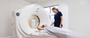 A health care professional in navy blue scrubs prepares a patient for a diagnostic imaging scan using a CT scanner in a hospital imaging department.
