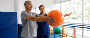 A rehabilitation therapist in navy blue scrubs guides a patient through a therapy exercise using a large exercise ball in a rehabilitation clinic.