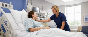 A health care professional in navy blue scrubs smiles while comforting a patient in a hospital bed during a bedside visit.