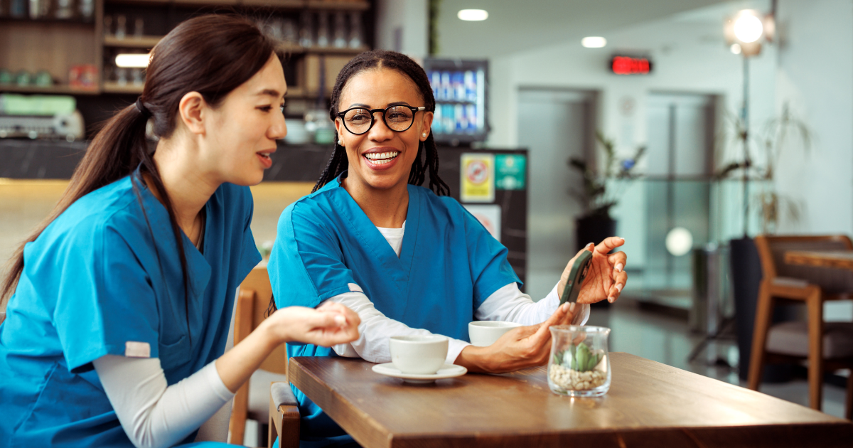 Two female clinical professionals talking in a cafeteria drinking coffee and looking at a phone