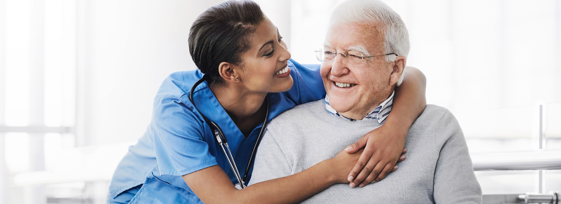 A smiling nurse in blue scrubs wraps her arms affectionately around an elderly man with white hair and glasses. They both smile at each other in a bright, welcoming health care setting.