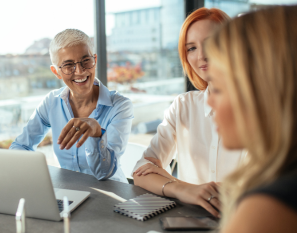 Three professional women sit at a table in front of a laptop, smiling and discussing something together. The setting is a modern office space with large windows and a city view in the background.