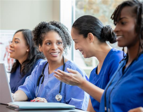 Four nurses in blue scrubs sit at a table engaged in conversation. One nurse smiles while another gestures as she speaks. A laptop and notebooks are visible, suggesting a team meeting or training session.