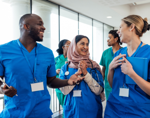 Three health care professionals in blue scrubs smile and laugh while talking together. One wears a stethoscope and another holds a blue folder. Additional teammates in scrubs are visible in the background near large windows.
