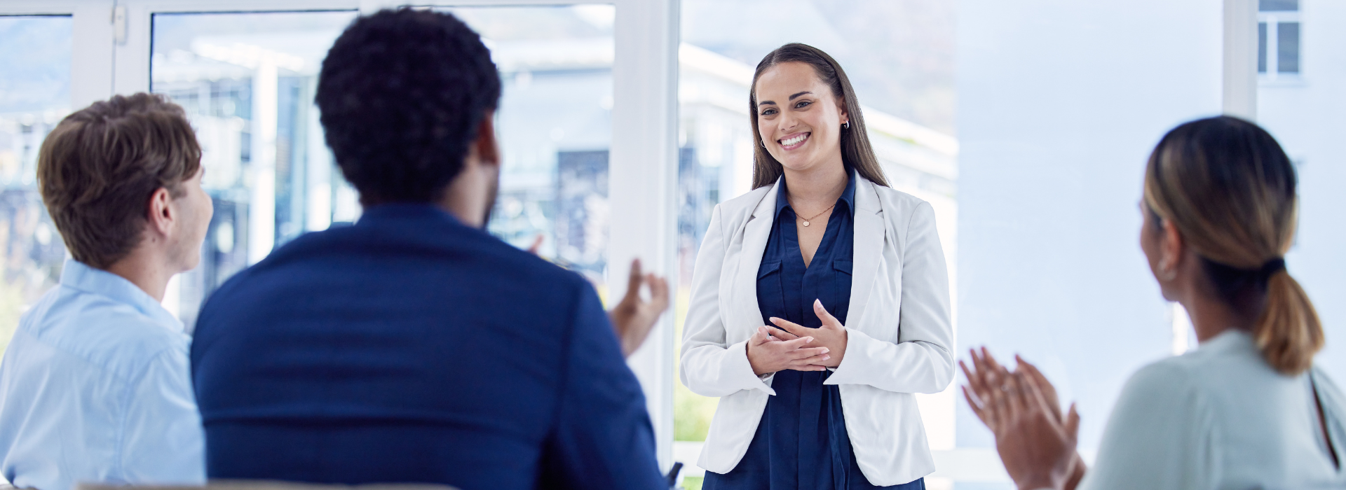 A smiling woman in a navy dress and white blazer stands and presents to a group of colleagues seated around a table. The room is filled with natural light from large windows and the group appears engaged and supportive.