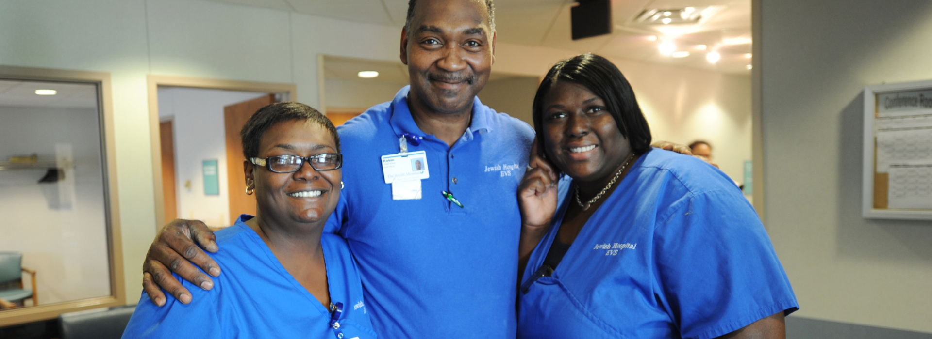 Two women and one man in matching blue EVS uniforms stand close together, smiling in a hospital hallway. The man’s badge reads “Jewish Hospital EVS,” and they appear to be part of a supportive, friendly team.