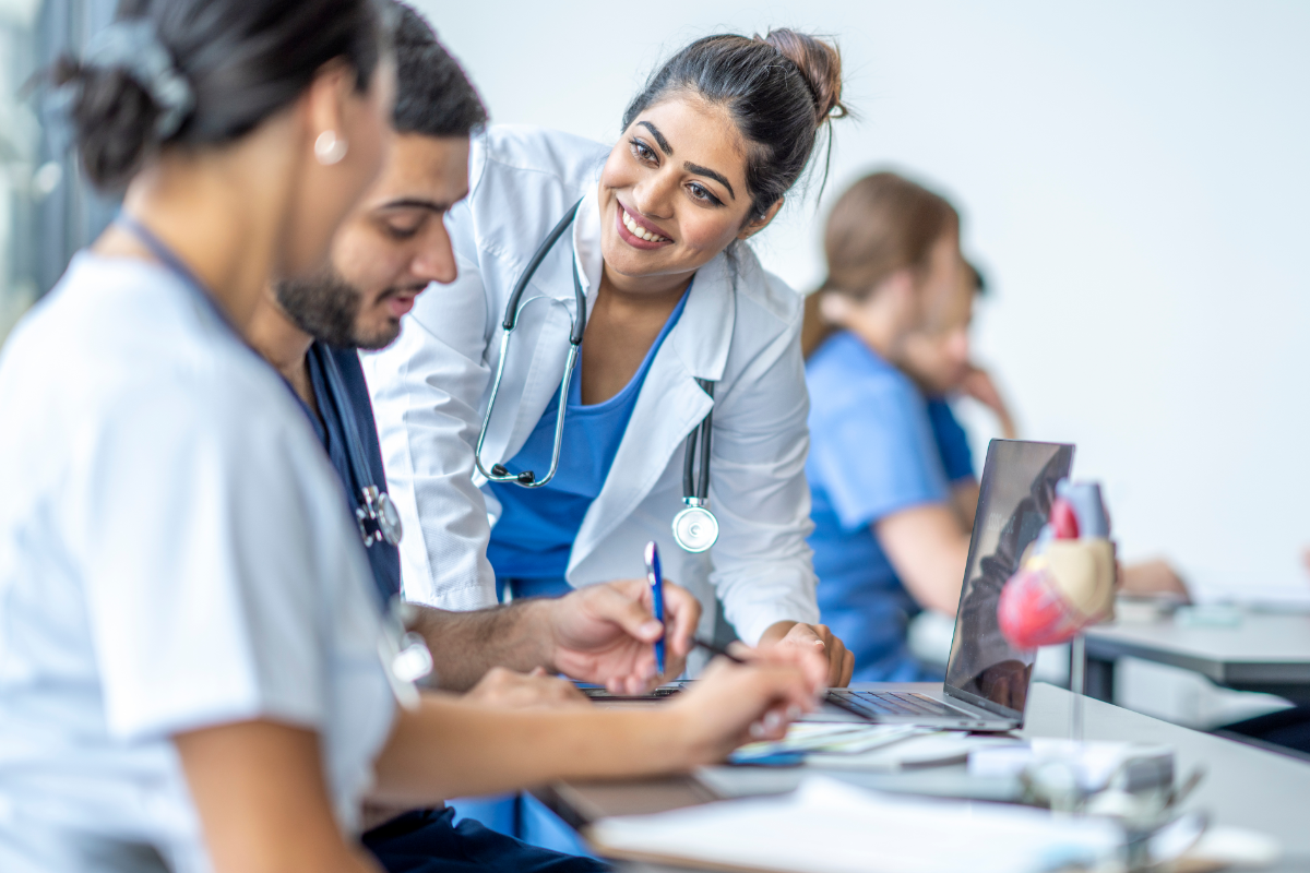 A group of health care students and a smiling instructor in a white coat engage in discussion around a laptop in a clinical classroom setting, with medical models and notepads on the table.