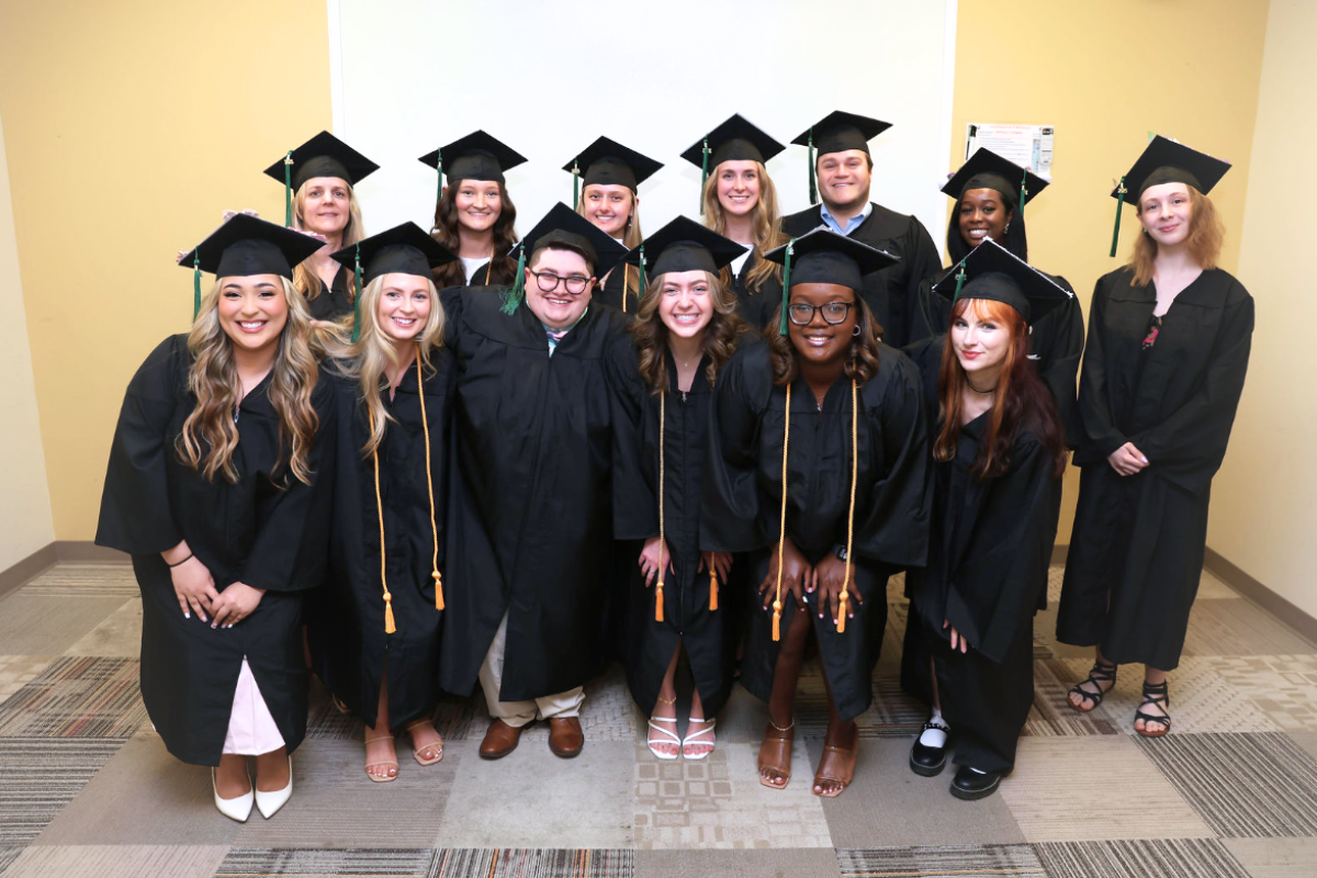 A diverse group of smiling graduates in black caps and gowns pose together for a celebratory photo. The front row kneels slightly while the back row stands, all radiating joy and accomplishment.