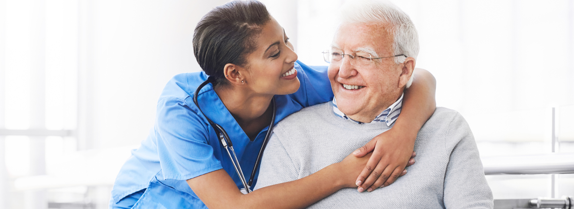 A smiling nurse in blue scrubs wraps her arms affectionately around an elderly man with white hair and glasses. They both smile at each other in a bright, welcoming health care setting.