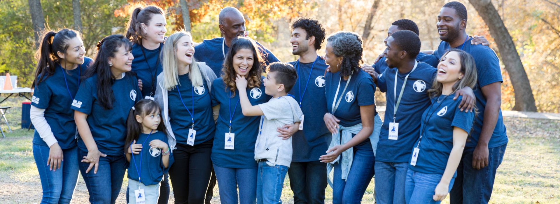 A large group of smiling volunteers stands close together outdoors in a park. They wear matching blue T-shirts with a circular logo and name badges, laughing and engaging with one another on a sunny day.