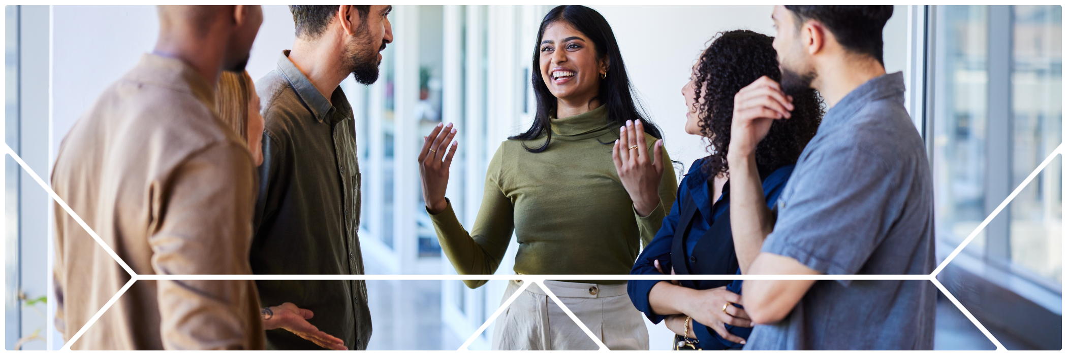 A diverse group of young professionals standing in a bright, modern office space, engaged in a friendly and animated conversation. One woman in the center is smiling and gesturing with her hands while others listen attentively