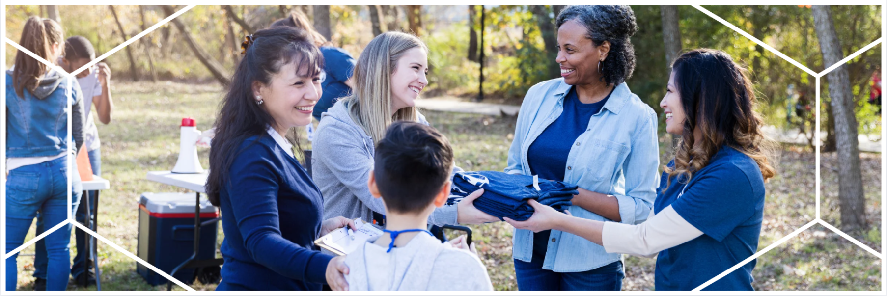 Four women and one child engage in a cheerful outdoor volunteer event, handing out clothing in a wooded park setting. The image is framed by Bon Secours Mercy Health’s signature geometric brand pattern.