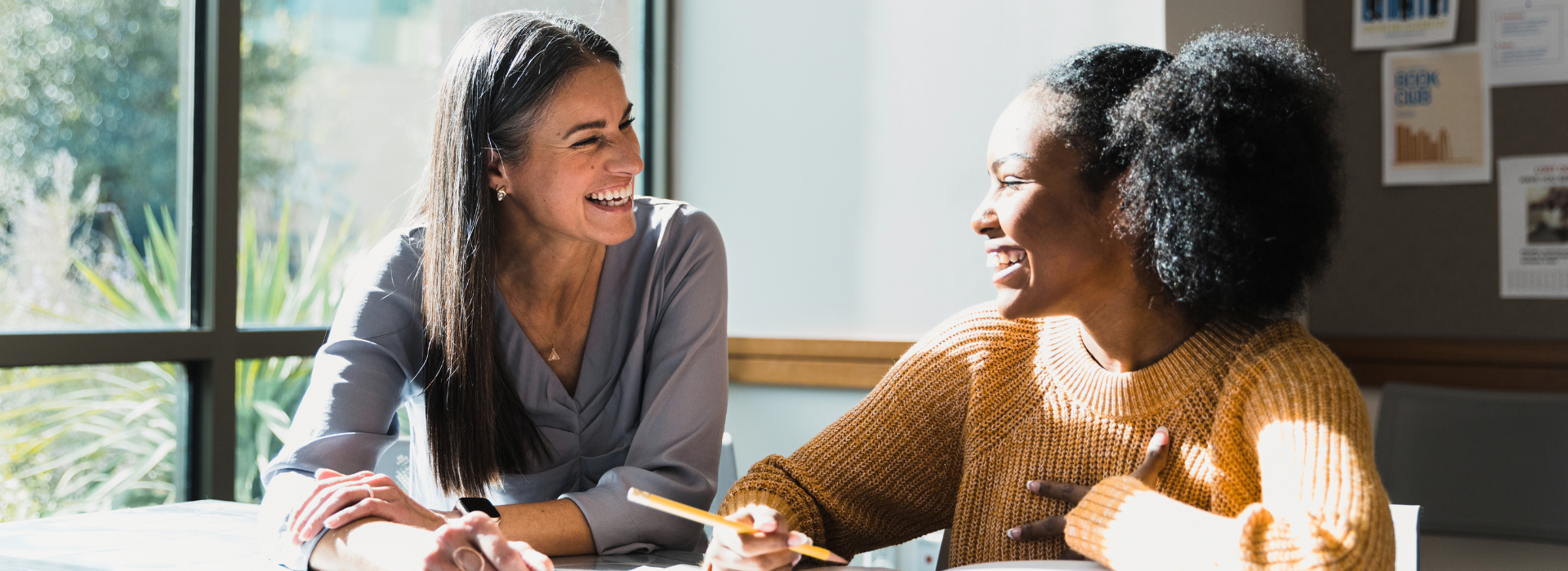 Two women sit at a table by a sunny window, smiling and talking. One wears a lavender blouse and the other an orange sweater, with papers and a pencil in front of them, suggesting collaboration or mentorship.
