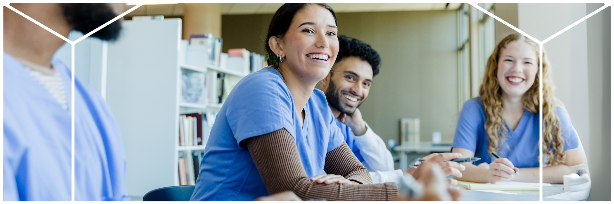 A group of diverse nursing or medical students in scrubs sit at a table in a bright classroom or library, smiling and engaged in discussion or study.