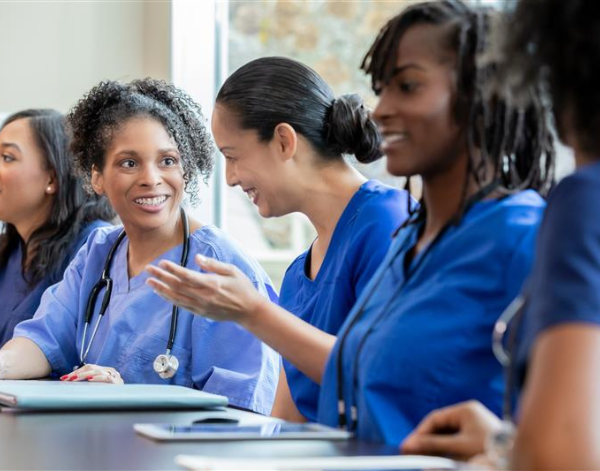 Four nurses in blue scrubs sit at a table engaged in conversation. One nurse smiles while another gestures as she speaks. A laptop and notebooks are visible, suggesting a team meeting or training session.
