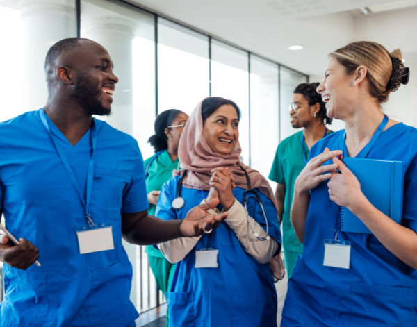 Health care workers in blue and green scrubs walk down a bright hallway, laughing and chatting with one another. The group includes individuals of various backgrounds, showcasing a welcoming team environment.