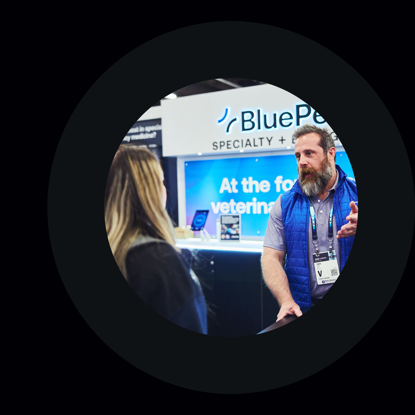 A BluePearl Associate with a beard and blue vest chats with a person with long blond hair at BluePearl's conference booth.