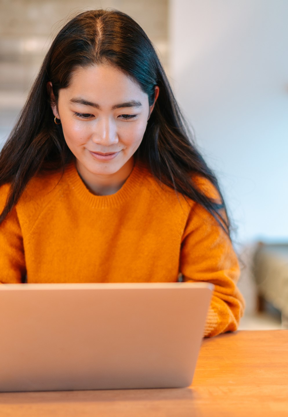 A woman in an orange sweater smiles while using her laptop.