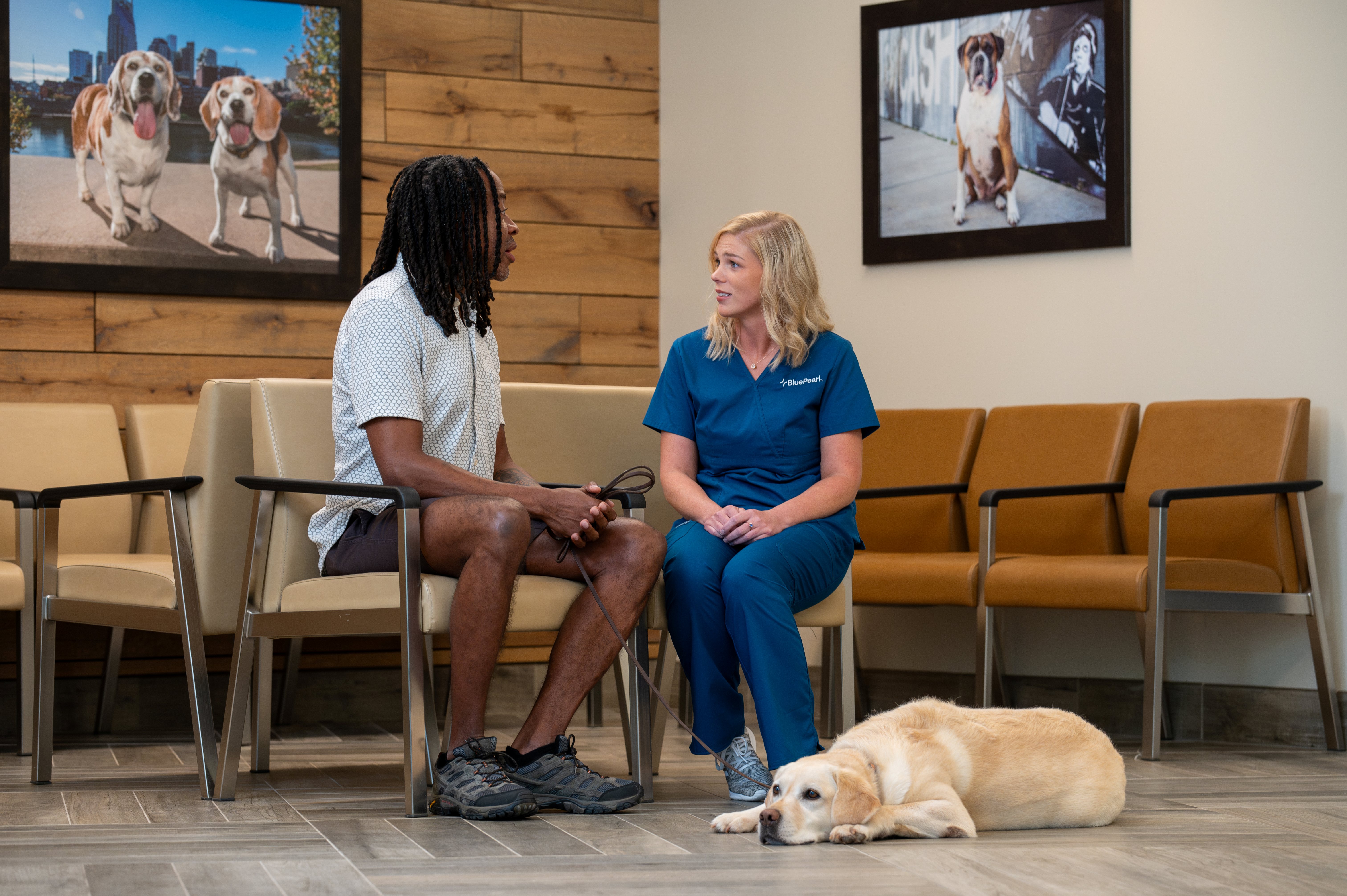 A man with long dark hair talks to a female BluePearl Associate while a yellow lab relaxes on the floor beside them.