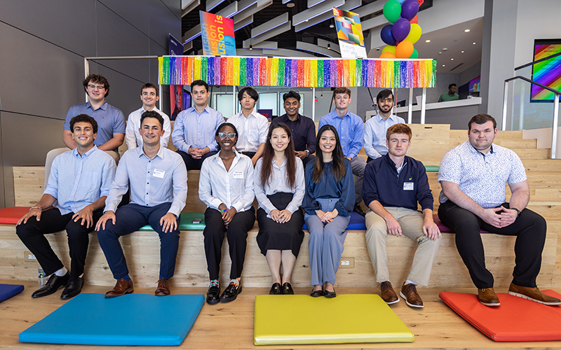 A group of Cencora interns are seated, smiling directly at the camera, in a modern light wood stadium style sitting area in the Cencora headquarters building. In front of them are brightly colored floor cushions. 