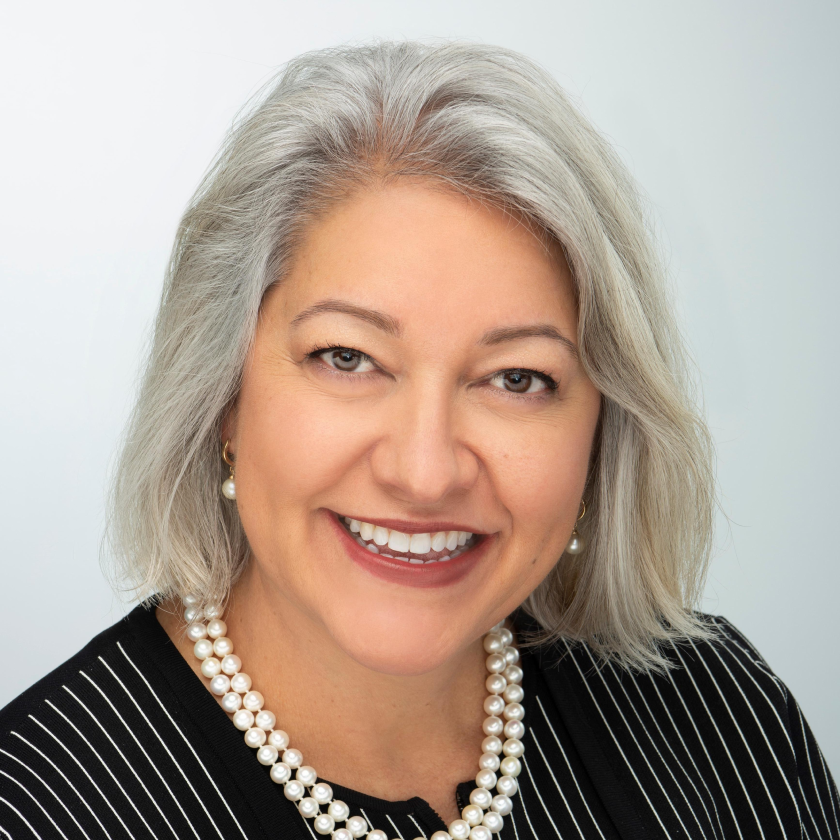 Photograph of Ann Anaya. Ann, a light-skinned person with gray, shoulder-length hair and brown eyes, is smiling at the camera. They wear a pearl necklace, earrings, and a black shirt with vertical white stripes. They are in front of a gray background.