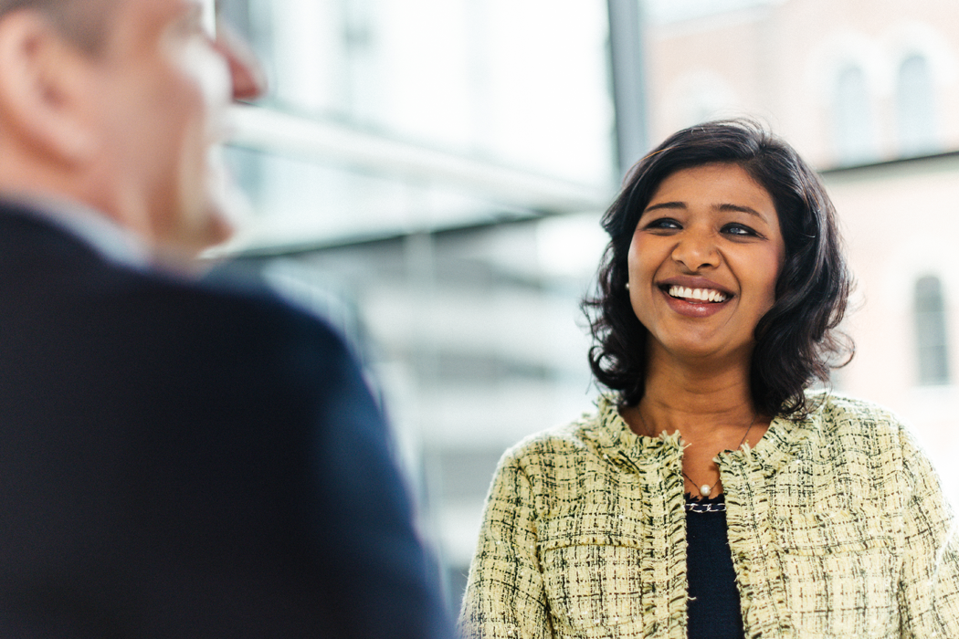 A Cencora team member in business attire is smiling and conversing with another person in a professional setting. They are standing in front of large windows.