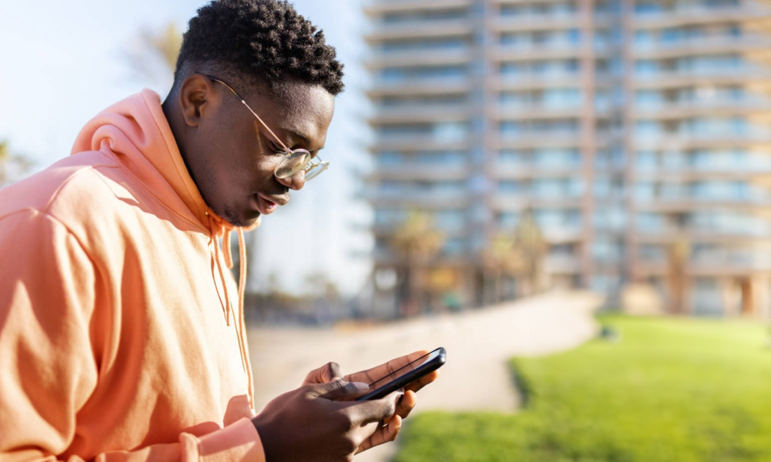 A dark-skinned young adult looking down at their cell phone. They have short hair and wear an orange hooded sweatshirt and glasses. They are outdoors in front of a large building.