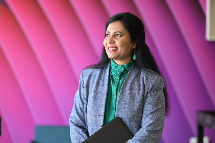 A Cencora team member stands in front of a colorful magenta wall. They smile and look off to their left - they are wearing business professional clothing - a green top and a grey business jacket.