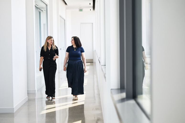 Two Cencora team members are talking while walking down a brightly, naturally lit hallway. The walls are white and there is sunlight coming in from the window. 