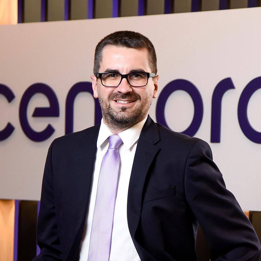 A portrait of JM, Sr Director of Shared Services at Cencora. Behind JM is the Cencora logo on the wall. JM wears a black business jacket, white shirt, and a tie.