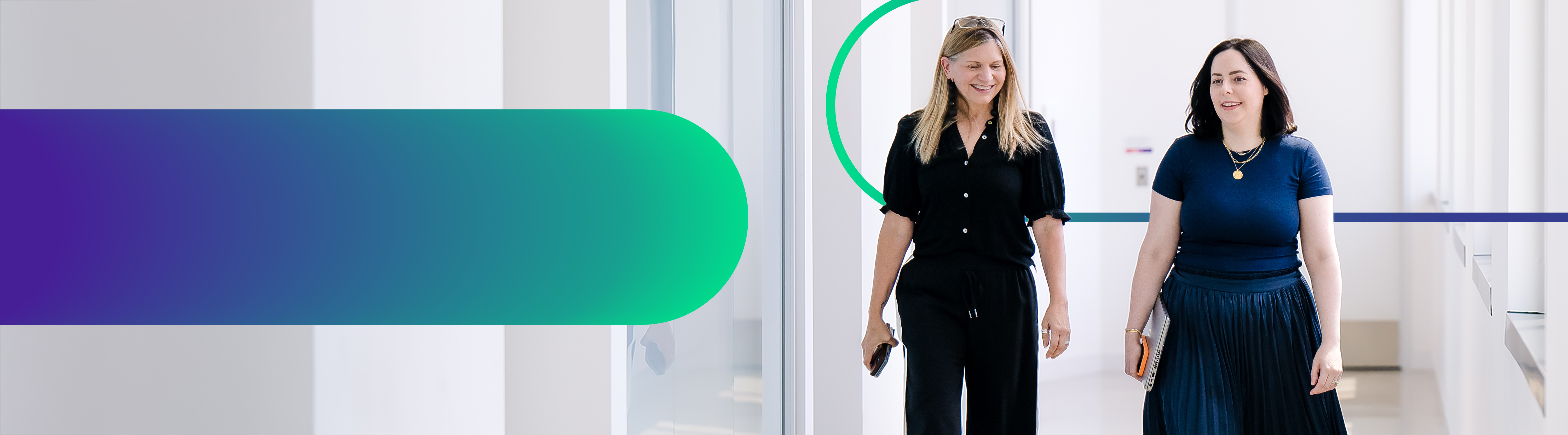 Two Cencora team members walk through a bright naturally lit hallway and are conversing. One team member holds a lap top and a phone, the other holds a phone. 