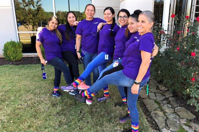 A group of WIN ERG members stand together with their arms around each other smiling for a photo. They are showing off their rainbow colored Cencora socks.