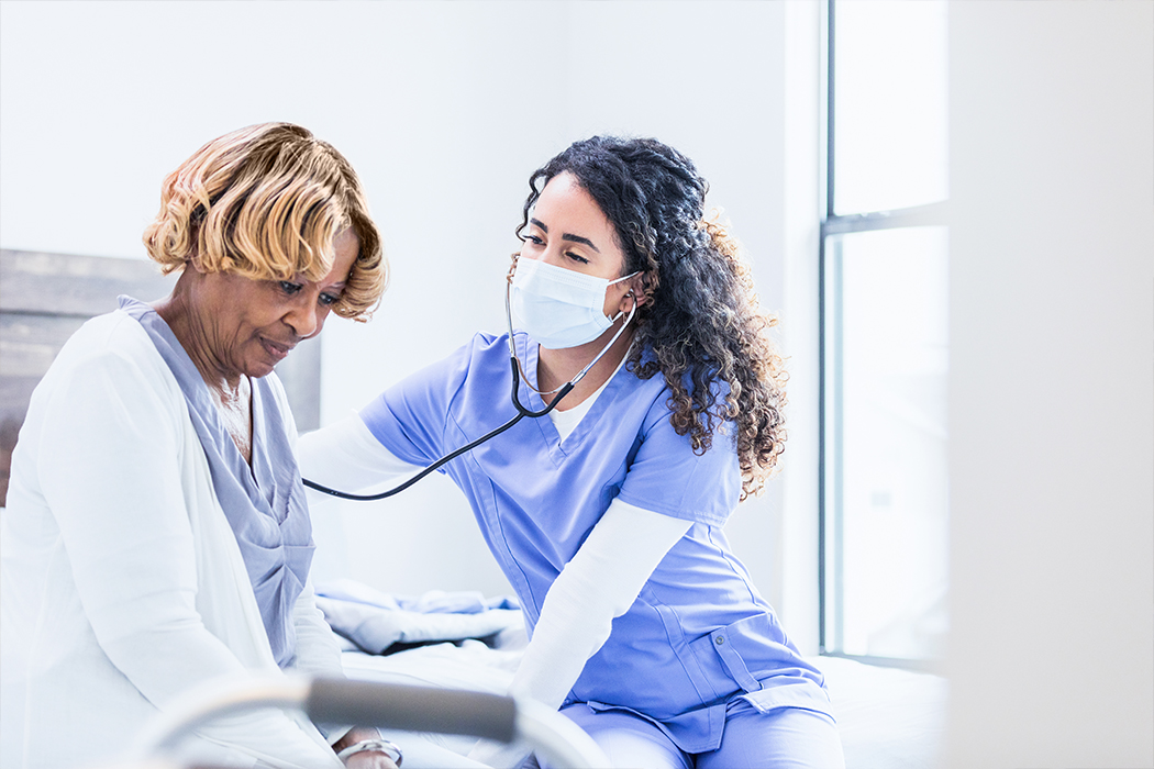 A nurse uses a stethoscope on a patient in a hospital setting. The nurse is wearing a protective face mask.