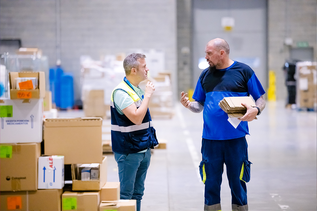 Two Cencora team members are in a distribution center. They are standing and talking. Next to them is a stack of packages ready for shipment.