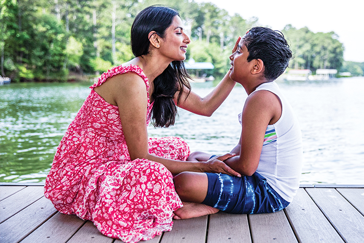 A dark-skinned parent and child sitting cross-legged on a dock next to a body of water. The parent wears a pink dress and the child wears blue shorts and a white tank top. The parent has a hand on the child’s face and they both smile.