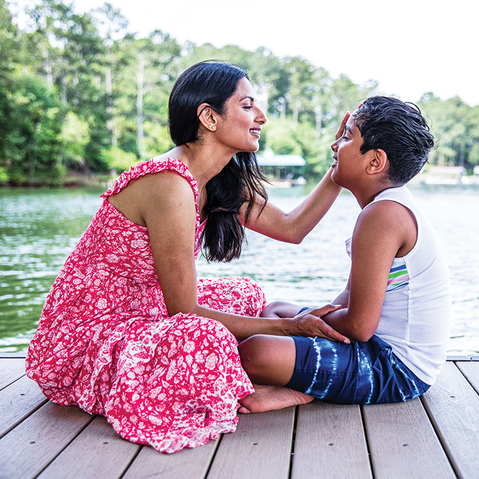 A dark-skinned parent and child sitting cross-legged on a dock next to a body of water. The parent wears a pink dress and the child wears blue shorts and a white tank top. The parent has a hand on the child’s face and they both smile.