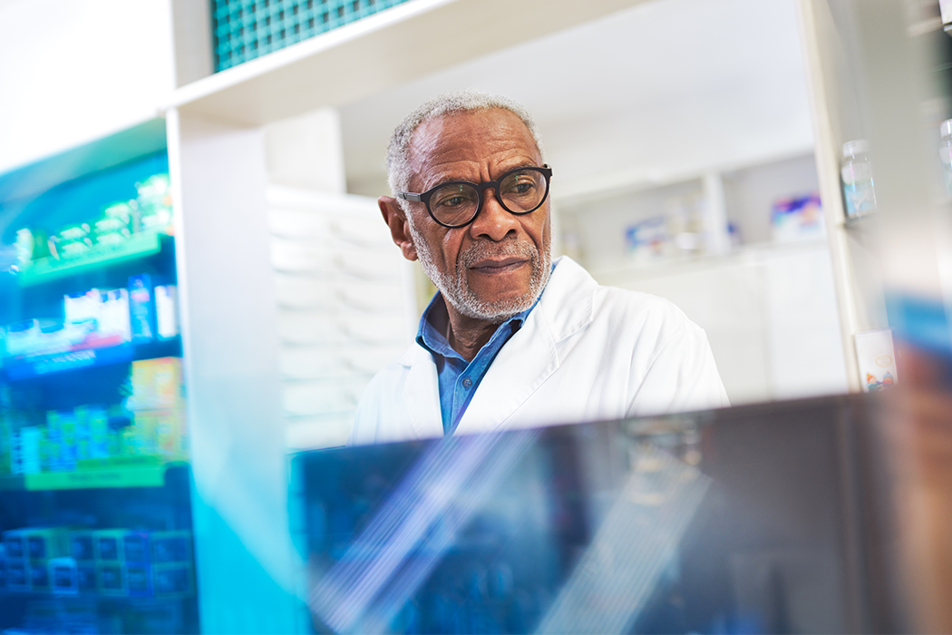 A dark-skinned pharmacist glances down at something out-of-frame as they work in a pharmacy. They wear a white lab coat and black glasses.