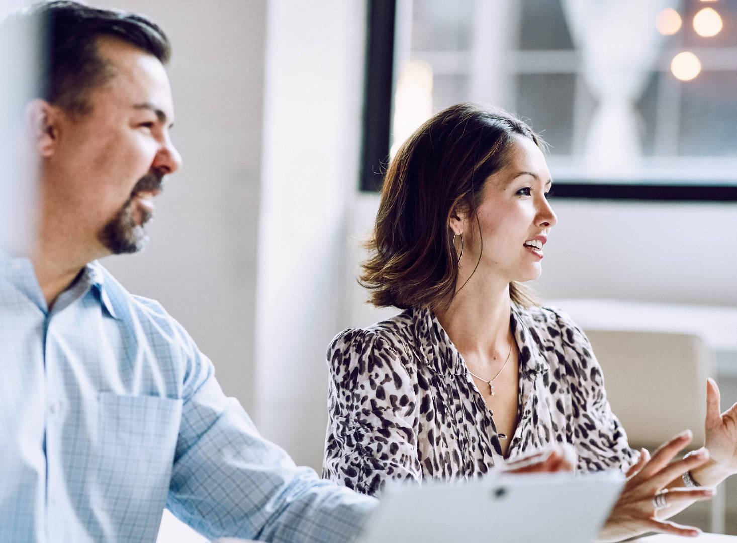 Two office workers sit side-by-side while looking at someone off-camera in a conference room setting.