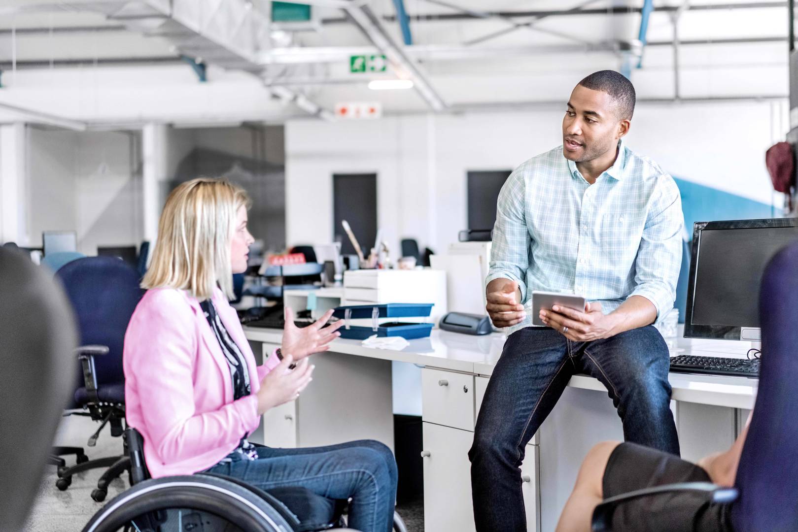 Two colleagues engage in discussion in an open office setting. On the left is a woman sitting in a wheelchair. On the right is a man sitting on top of a desk.