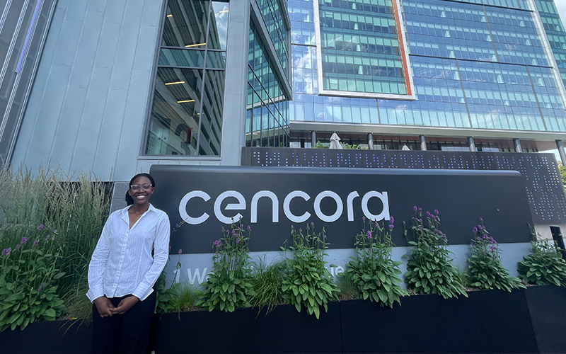 A Cencora intern stands outside of the Cencora Headquarters building. They are standing in front of a sign with Cencora's logo on it - behind them is Cencora's HQ - a tall glass building with many windows.