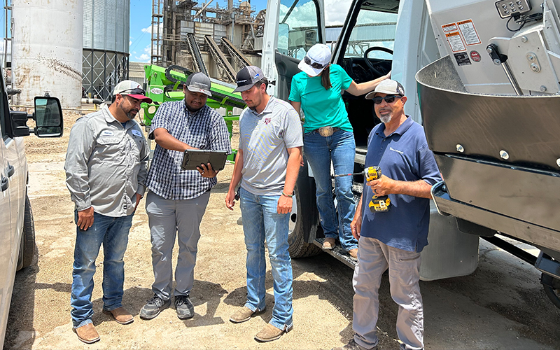 An intern meets with a group of 4 team members out in the field. They are standing in the parking lot of an industrial park and are looking at a tablet. 