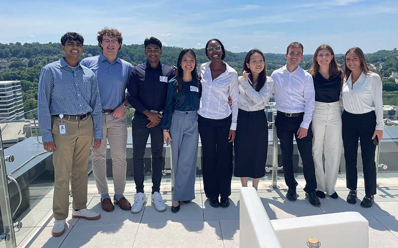 A group of 9 Cencora interns pose for a photo up on the rooftop terrace of Cencora's headquarters building. They are dressed in professional attire and are smiling while looking at the camera.