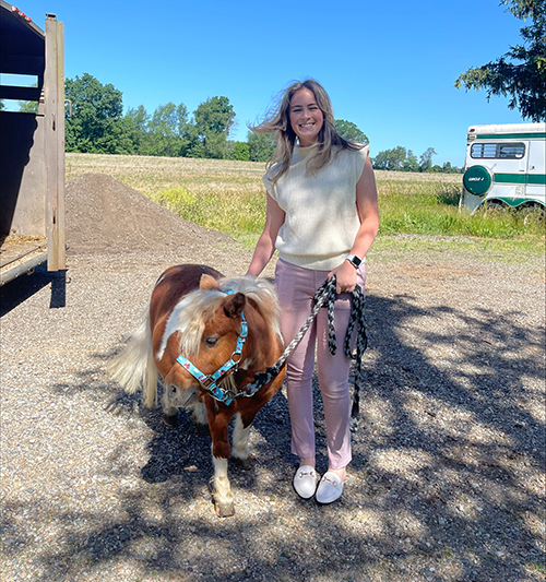 A Cencora intern smiles as they hold the reigns to a mini horse. The intern is smiling and the horse is looking off to the side. They are in an outdoor setting in a field.